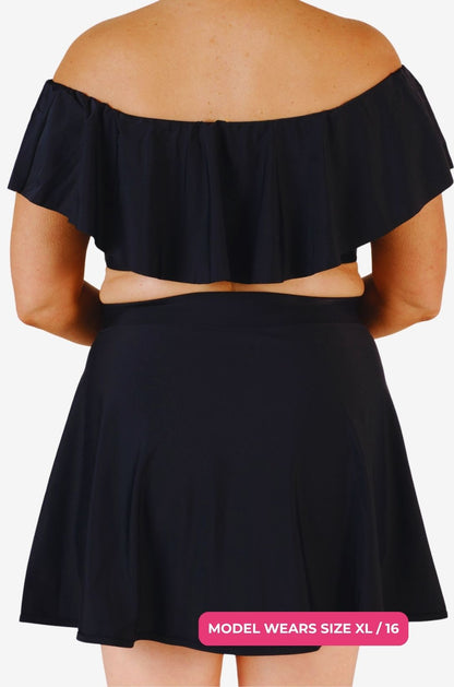 Back view of a black off-shoulder swim top and high-waisted swim skirt worn by a model size XL/16 set on a grey background