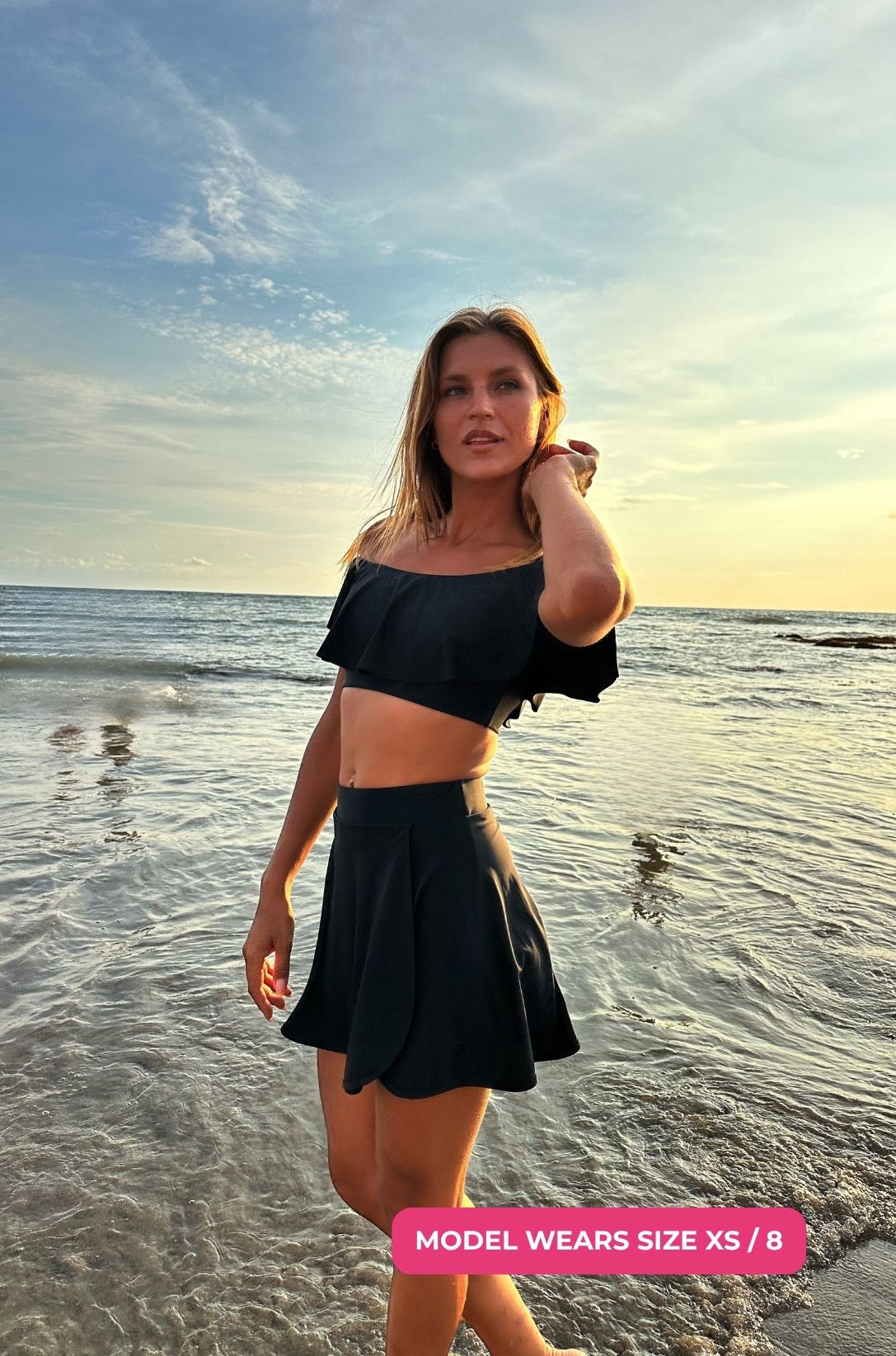 Woman size XS/8 in black off-shoulder swim top and high-waisted swim skirt standing on a beach with ocean and sky in the background.