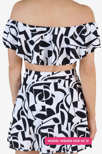 Back view of a black and white geometric patterned off-shoulder swim top and matching high-waisted swim skirt worn on a model size M/12