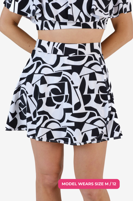 Close up view of a model size M/12 wearing a black and white geometric high-waisted swim skirt holding her hands behind her back on a grey background
