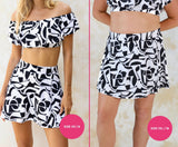 Two models wearing black and white geometric high-waisted swim skirts and matching off-shoulder swim tops with size XS/8 and 1XL/18 labels on a light cream background